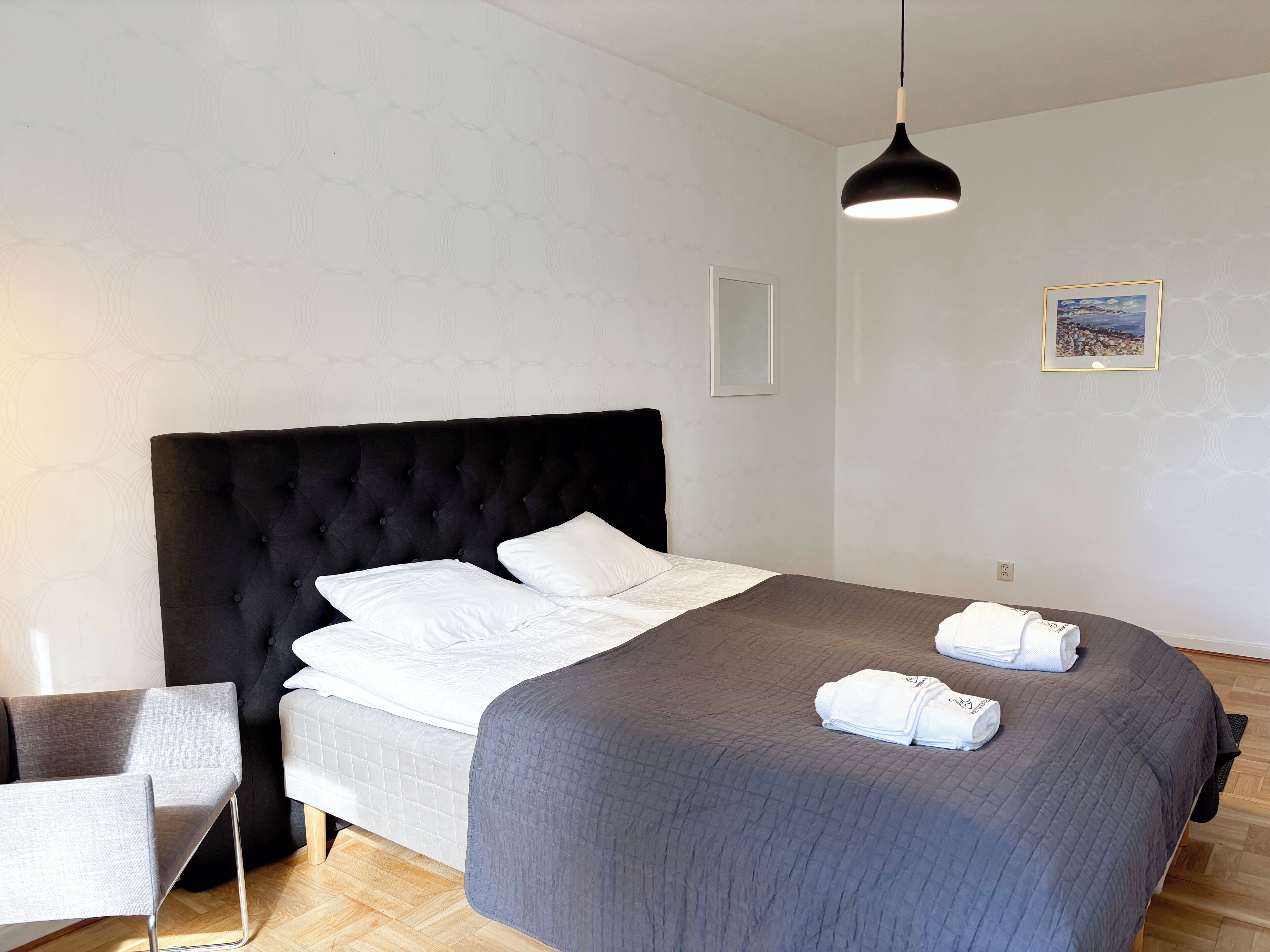 Una cama bien hecha con edredón gris, cabecera negra y dos almohadas blancas. Hay una silla gris al lado de la cama, una lámpara colgante negra arriba y un cuadro en la pared.