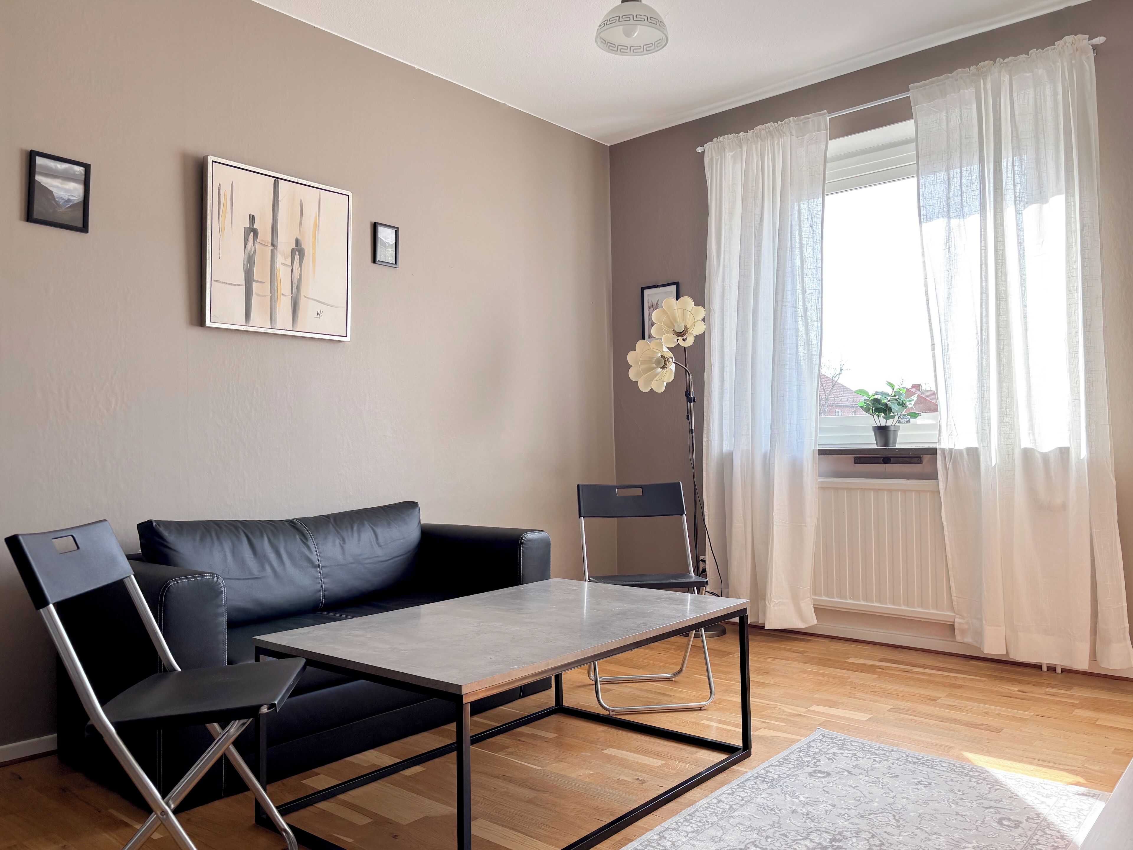Un salon moderne avec un canapé en cuir noir, une chaise pliante, une petite table et un lampadaire. Une fenêtre, ornée de rideaux légers, offre une vue sur l’extérieur.