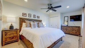 Quarto com cama de madeira, mesa de cabeceira, luminária, ventilador de teto, televisão e cômoda.