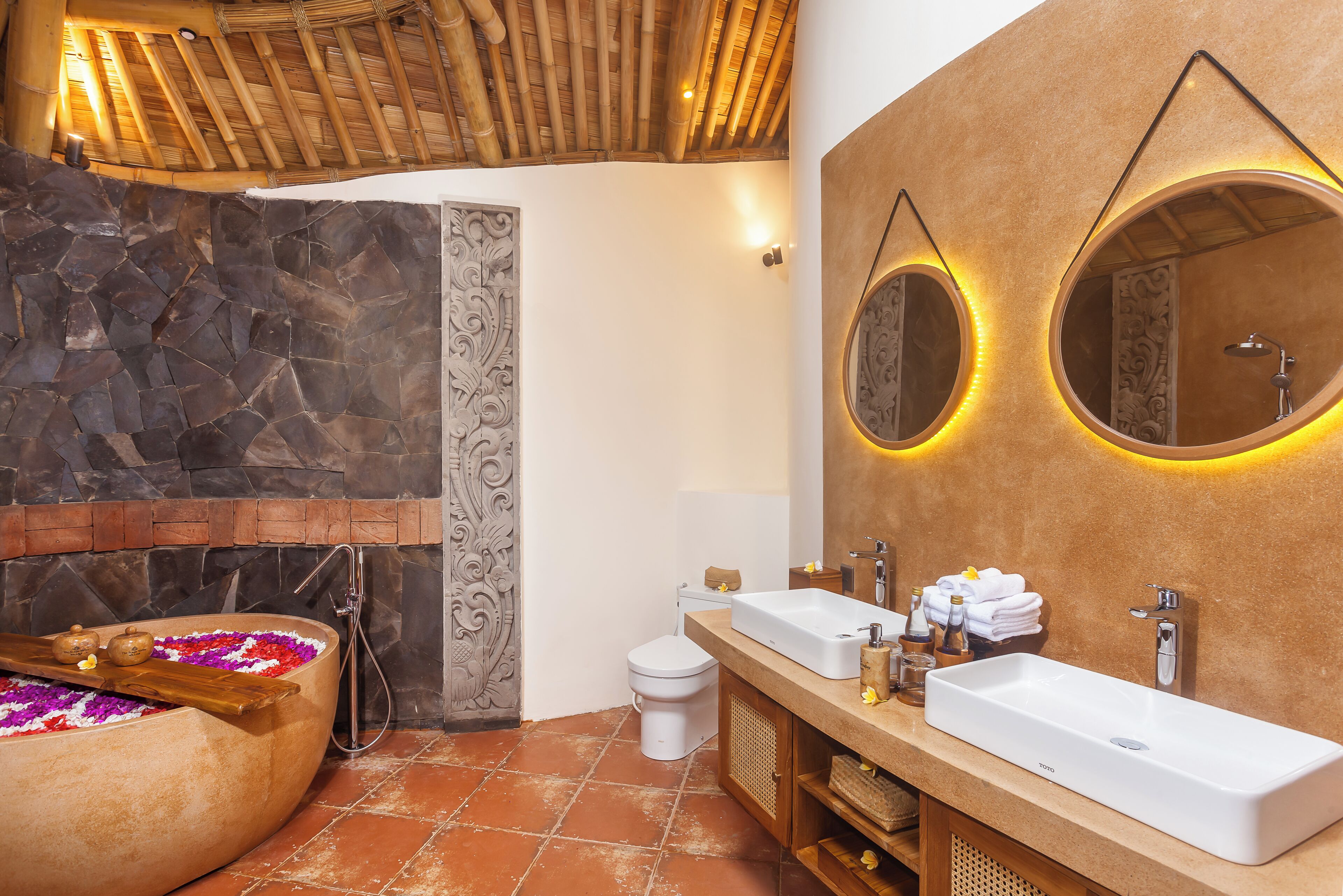 Een badkamer met een stenen muur, een bad met bloemen, een toilet en een wastafel met spiegel.