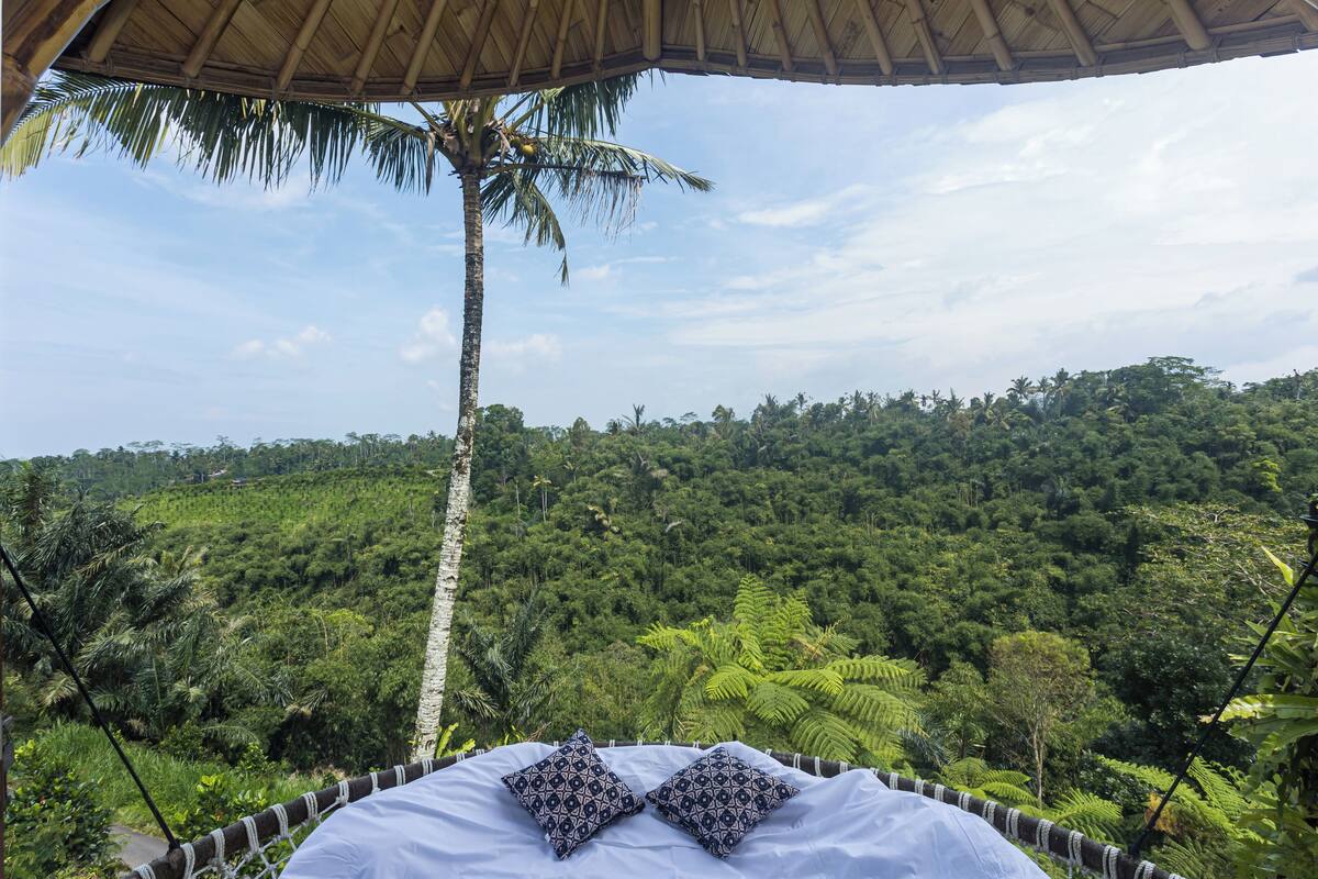 A bed with a white sheet and patterned pillows, set against a backdrop of lush greenery and a clear sky.