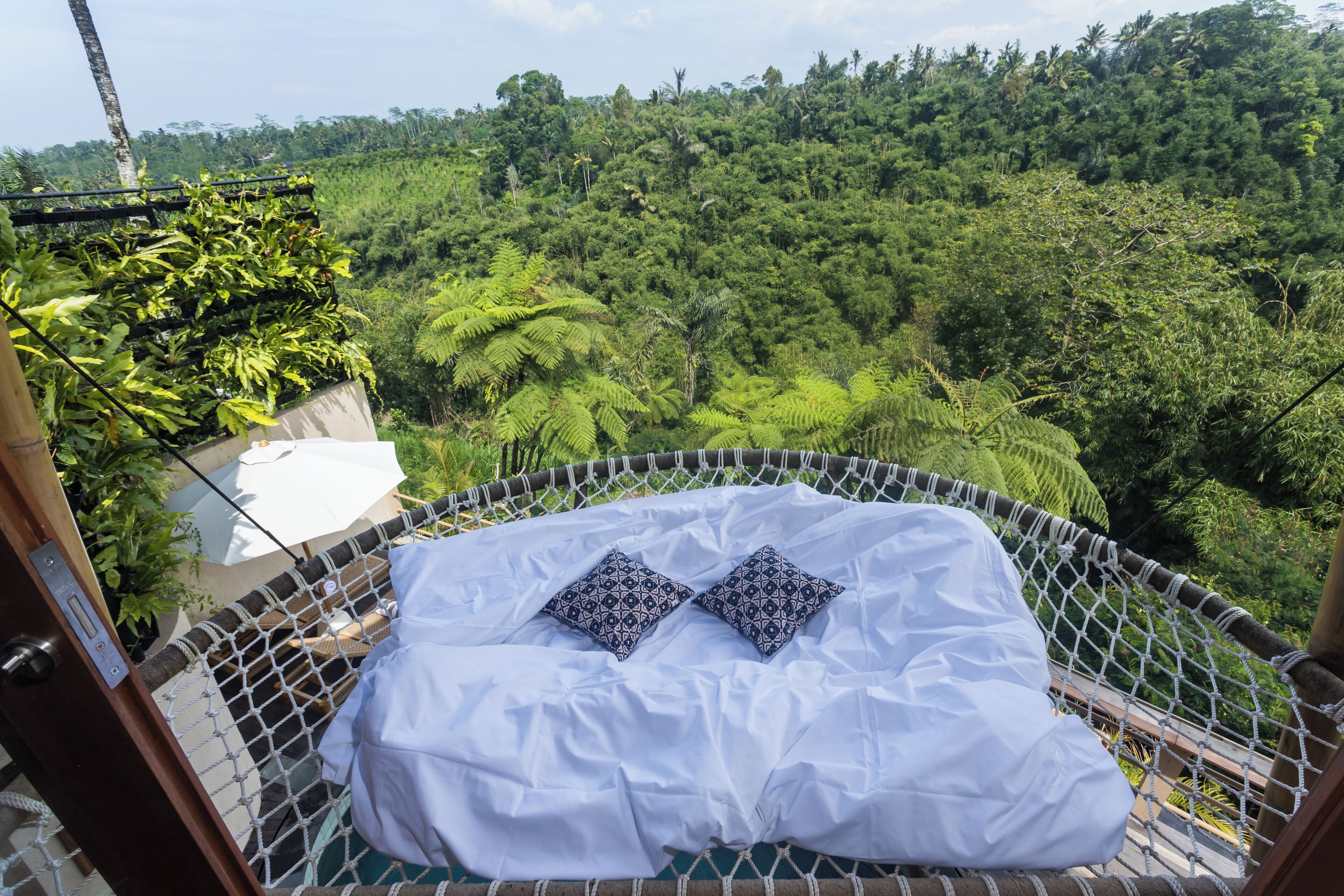 A hammock bed with white sheets and patterned pillows, set against a lush forest backdrop.