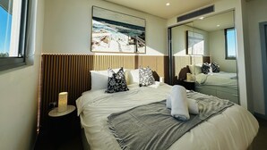 Una habitación de hotel moderna con una cama grande, una lámpara de noche, un cuadro de playa y un espejo amplio.