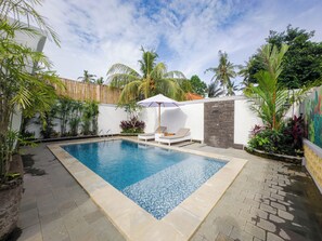 A rectangular swimming pool with a tiled edge, surrounded by a paved patio. There are lounge chairs and a white umbrella providing shade.