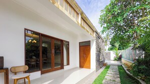 A modern house with a large sliding glass door, a small outdoor seating area, and a narrow pathway with grass and stone paving. - Pondok Merta-Two Bedroom Pool Villa Ubud (Ubud)