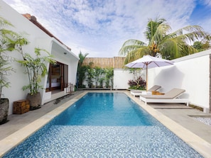 Uma piscina com piso de azulejos, paredes brancas e um deck de madeira. Em segundo plano, espreguiçadeiras e um cercado de madeira.