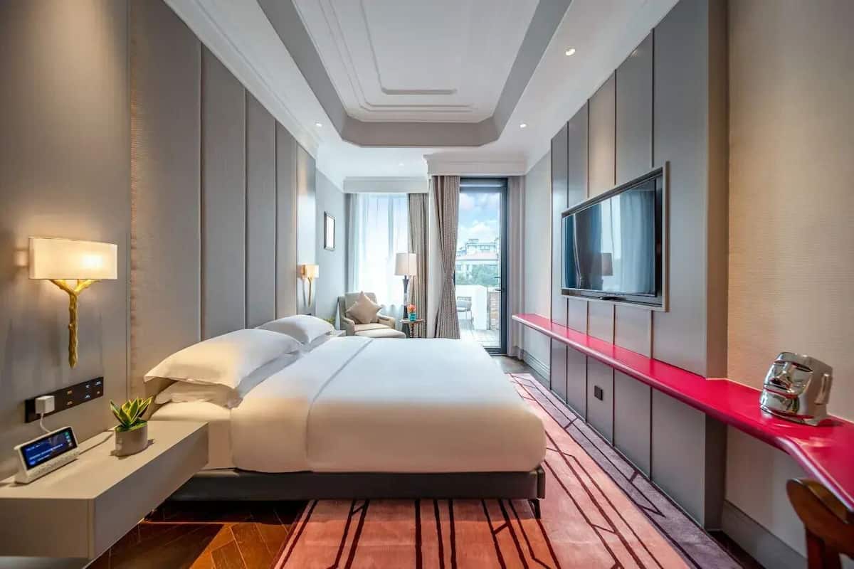 A modern hotel room with a large bed, a TV, a bedside table with a lamp, and a red counter.