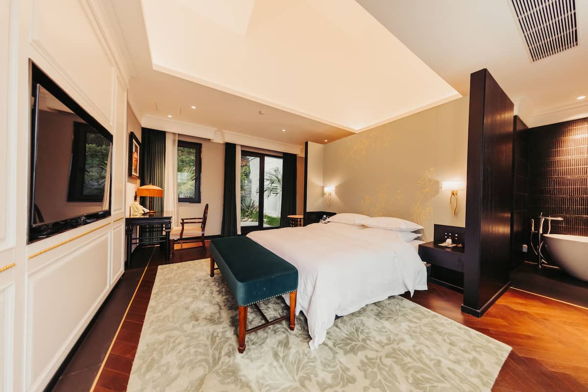 A modern hotel room with a large bed, a bench, a TV, a desk, and a bathroom with a bathtub.