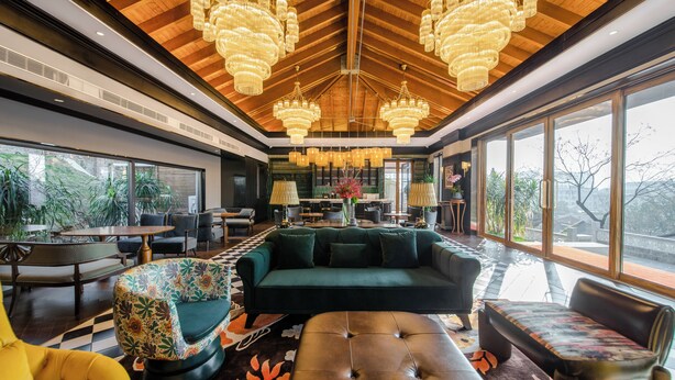 A spacious living area with a large green sofa, a tufted ottoman, and a colorful armchair. There are multiple tables and chairs, and the room features large windows with a view of greenery outside. - Seclusion with the mountain (Wuhan)