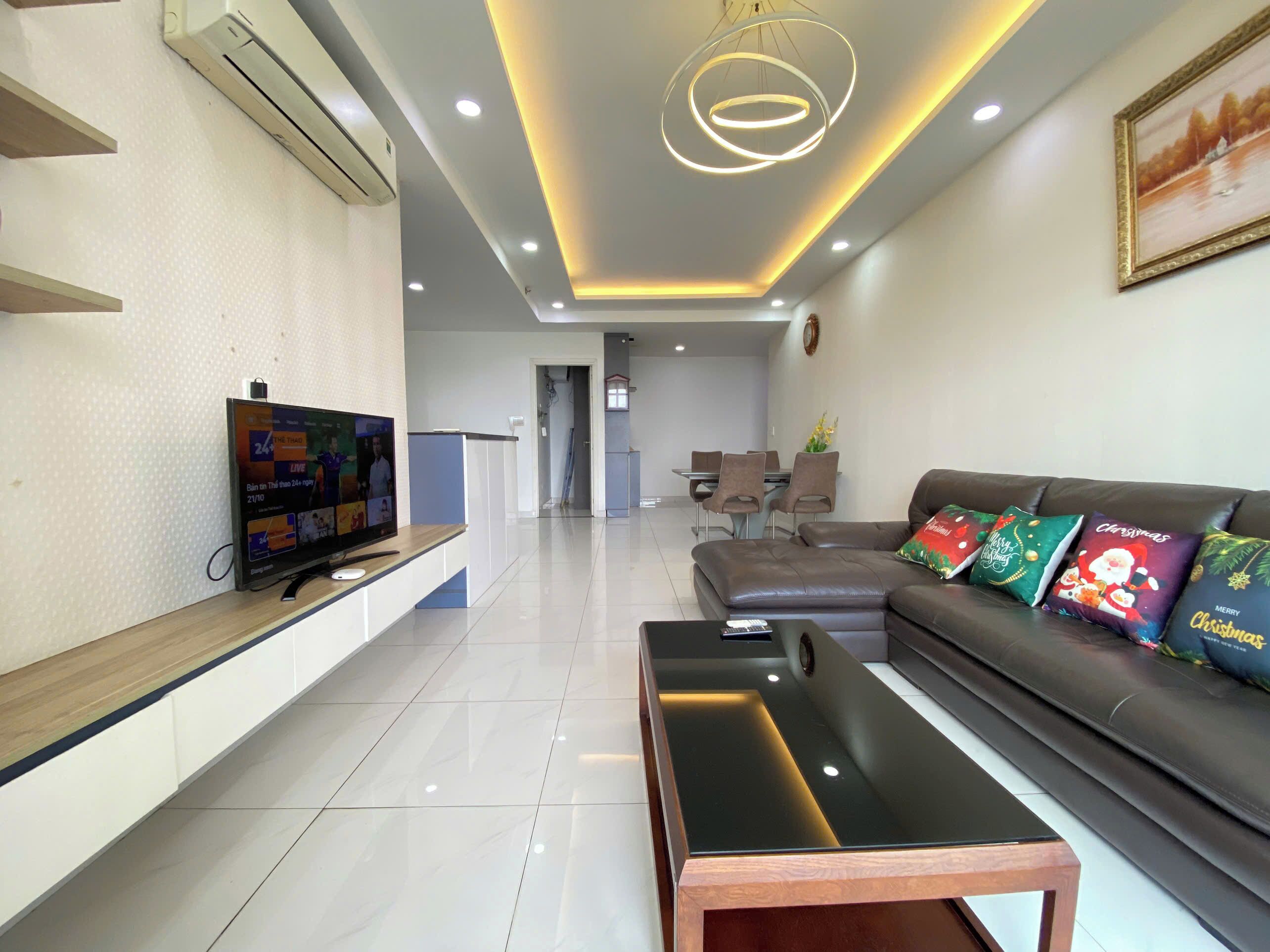 A modern living room with a flat-screen TV, a sofa with Christmas-themed cushions, a coffee table, and a dining area in the background.