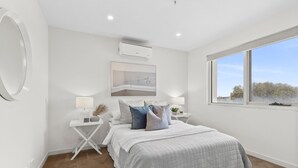 A bedroom with a bed, bedside tables, a mirror, an air conditioning unit, and a window with a view. - Walk to Beach and shops, Spacious, Ocean Views (Ocean Grove)