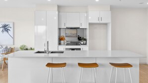 A modern kitchen with white cabinets, a central island with stools, and a painting of a palm tree on the wall. - Walk to Beach and shops, Spacious, Ocean Views (Ocean Grove)