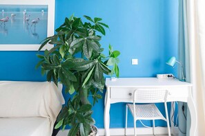 Ein Zimmer mit blauer Wand, einem weißen Schreibtisch mit Stuhl, einer Topfpflanze und einem Bilderrahmen mit Flamingos.