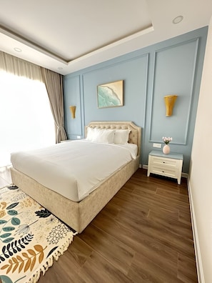 Un dormitorio con una cama grande, una mesita de noche, una lámpara de pared y un ventanal con cortinas.