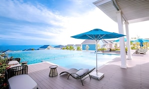 Zona de piscina con sillones, una mesa y sombrillas, con vista al océano azul y los edificios del complejo.