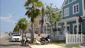 A street with parked cars and motorcycles, a white picket fence, and a blue building with white trim.