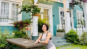 A woman sitting at a table outside a house with a porch, surrounded by greenery and flowers.