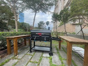 A barbecue grill with a metal cart, two wooden tables, and a stone-paved patio surrounded by greenery and buildings. - Sea ya inn (Hualien City)