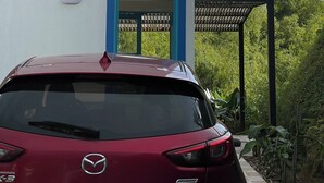 Un coche Mazda rojo estacionado frente a un edificio blanco con una puerta azul y una pérgola.