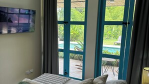 Un dormitorio con cama, televisor montado en la pared y vistas a una piscina y follaje a través de puertas azules.