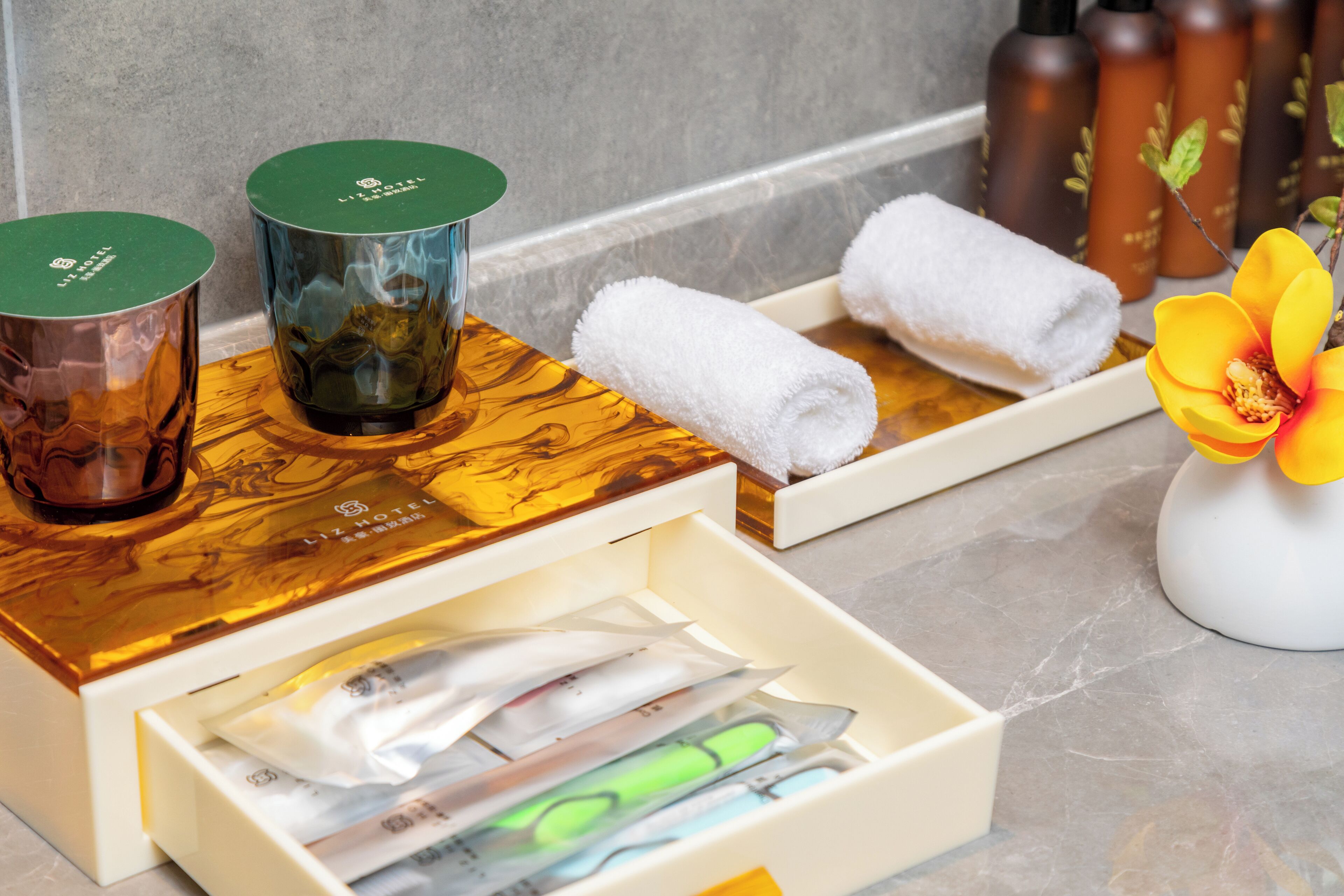 A bathroom with a wooden vanity, a mirror, and toiletries including a bottle of shampoo, a towel, and a flower vase.