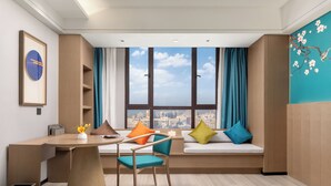 Une chambre d’hôtel moderne avec un bureau en bois, une chaise, un canapé agrémenté de coussins colorés, une grande fenêtre donnant sur la ville et une étagère fixée au mur.