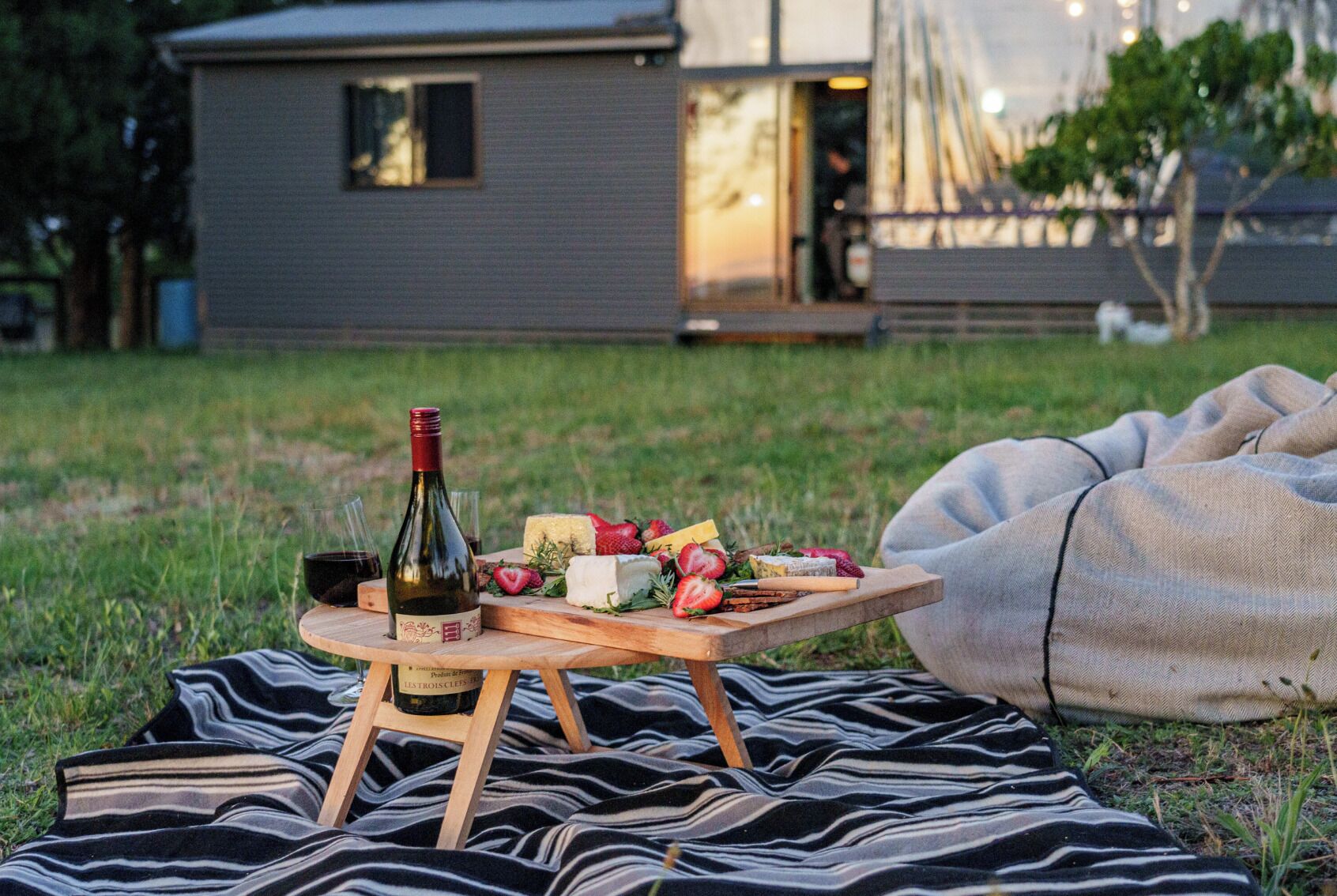A picnic setup on a grassy lawn with a bottle of wine, cheese, strawberries, and a wooden table.