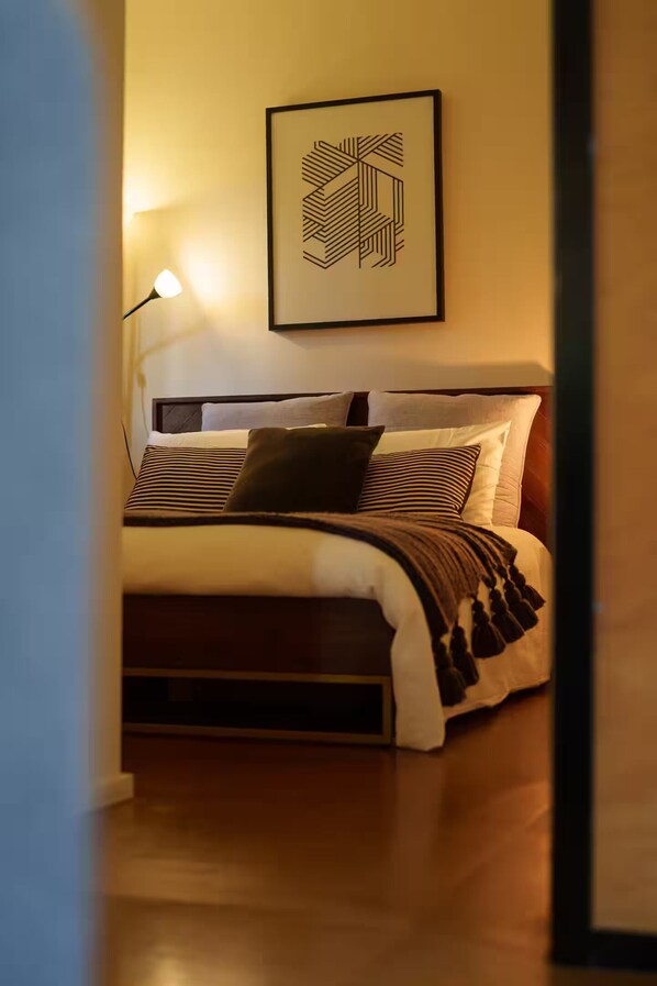 Ein ordentlich bezogenes Bett mit Streifenmuster, ein geometrisches Wandbild und eine Nachttischlampe.