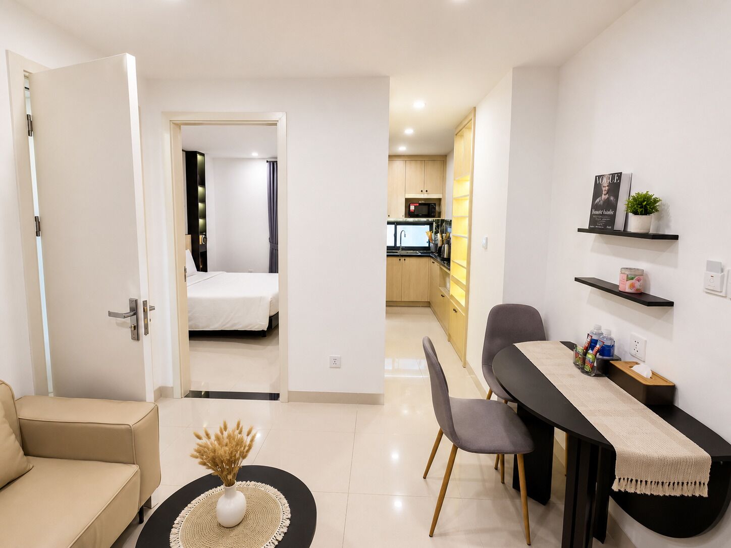 A modern hotel room with a bed, a dining area with a table and chairs, a kitchenette, and a bathroom visible through an open door.
