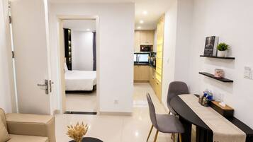A modern hotel room with a bed, a dining area with a table and chairs, a kitchenette, and a bathroom visible through an open door.