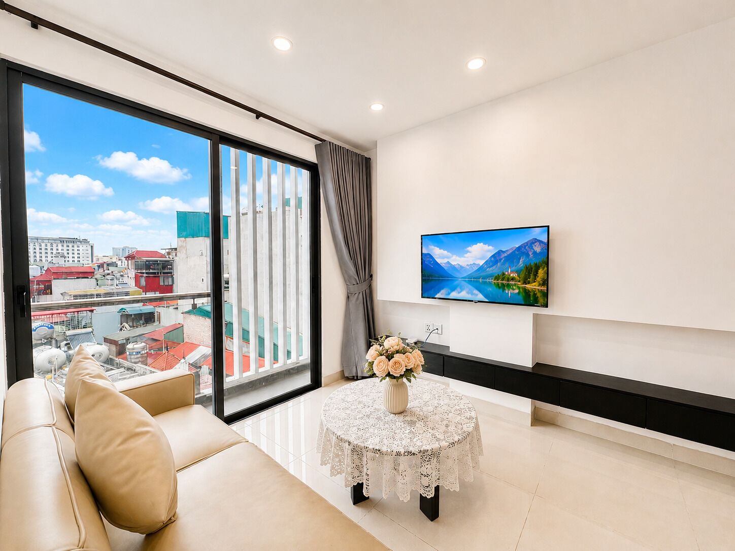 A modern living room with a sofa, a table with a vase of flowers, a flat-screen TV on the wall, and a large window with a cityscape view.