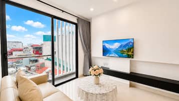A modern living room with a sofa, a table with a vase of flowers, a flat-screen TV on the wall, and a large window with a cityscape view.