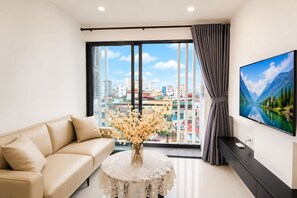 A modern living room with a sofa, a table with a vase of flowers, a large window with a city view, and a mounted TV. - Sun Residences Hotel and Apartment (Hanoi)