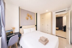 Habitación de hotel moderna con una cama, un escritorio con una computadora portátil, un armario y un baño visible a través de una puerta abierta.