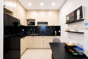Una cocina moderna con encimeras negras, armarios de madera, un microondas y un refrigerador. En la pared hay revistas enmarcadas y una pequeña estantería con botellas.
