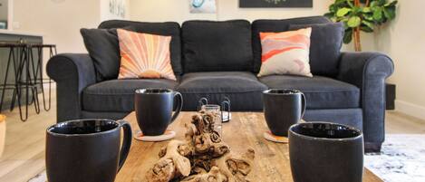 A living room with a dark blue sofa, two black coffee mugs on coasters, and a wooden table with a decorative centerpiece.