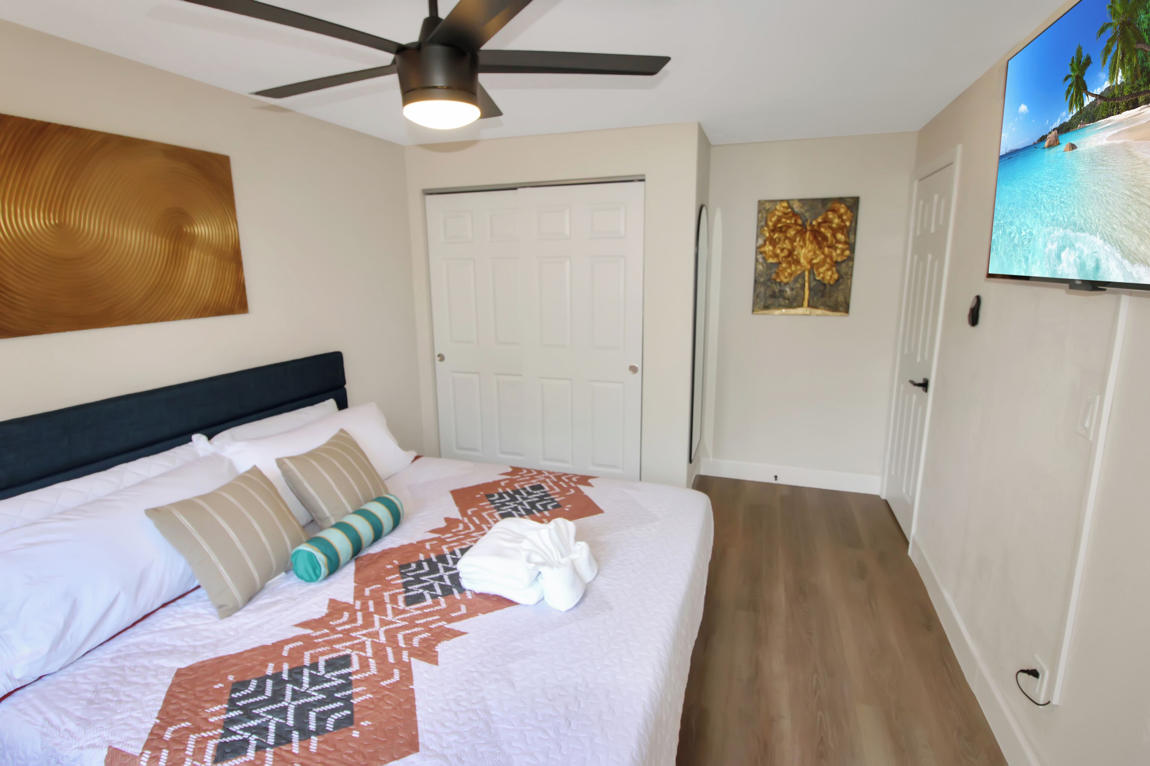 A neatly made bed with a patterned bedspread, a ceiling fan, and a television on the wall.