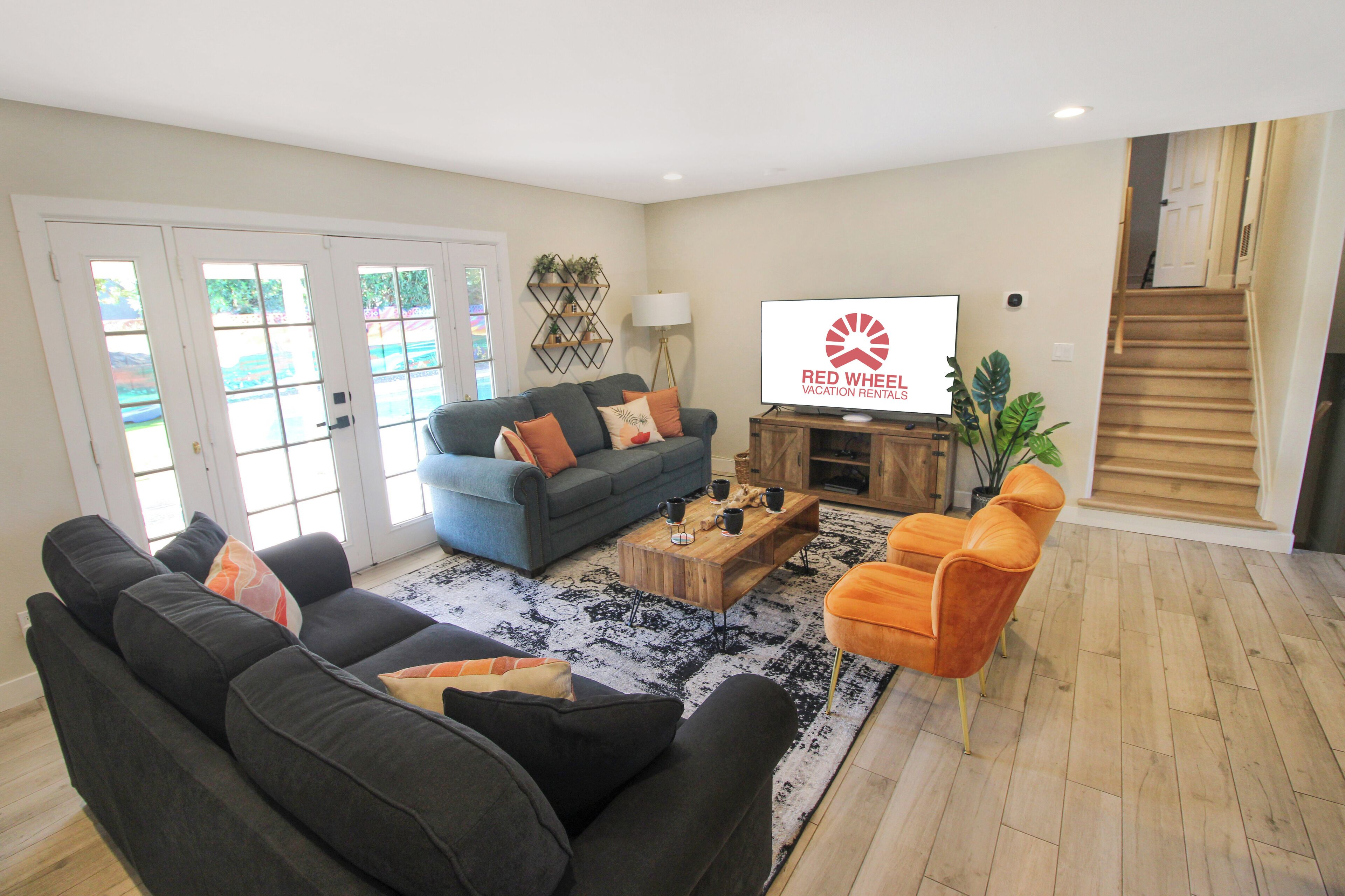 A living room with a grey sofa, a blue sofa, and an orange chair. There is a coffee table, a TV, and a staircase in the background.