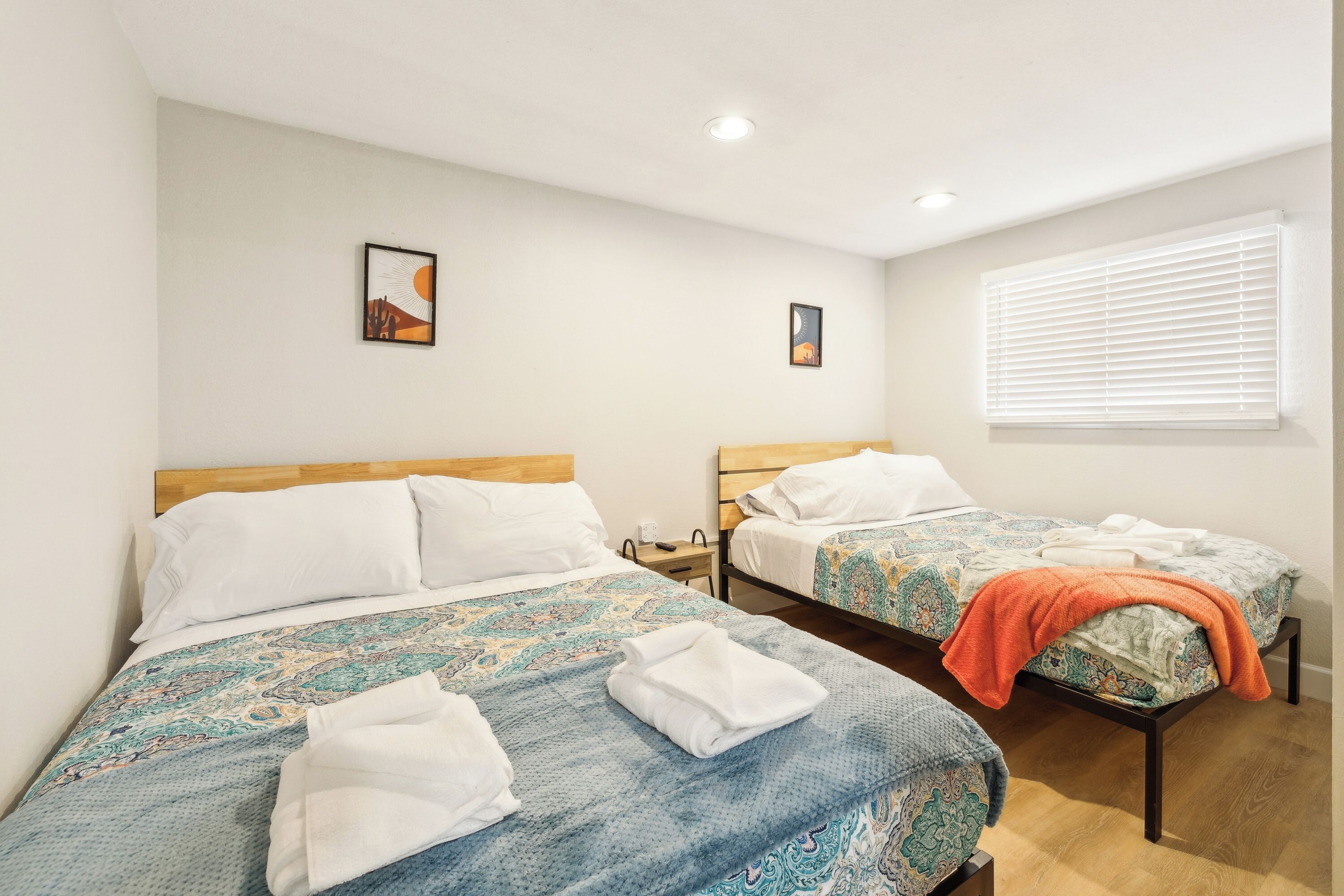 A hotel room with two single beds, each with a patterned bedspread and pillows. There are two wall-mounted nightstands with lamps, and a window with blinds.