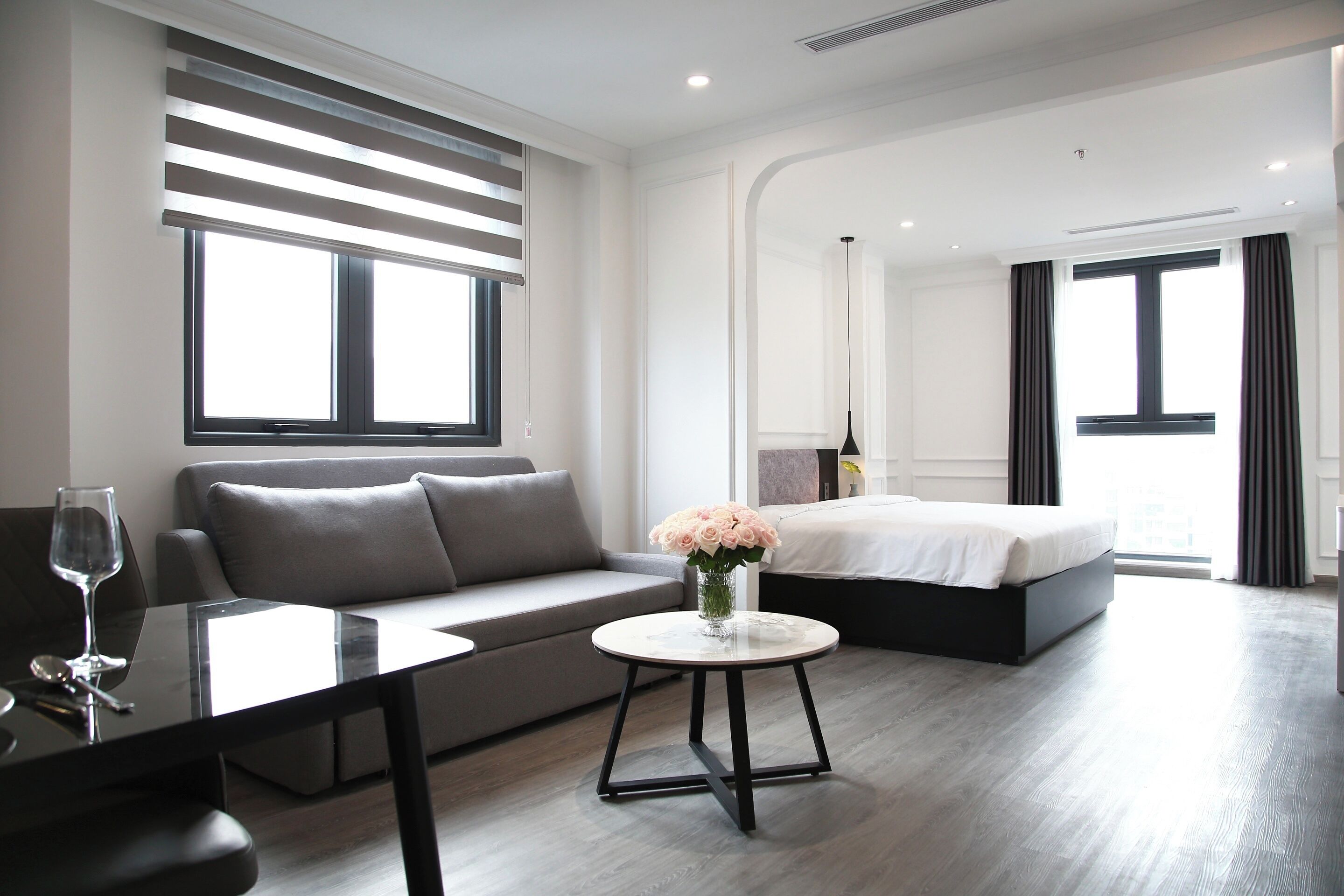 A modern hotel room with a sofa, a round table with a vase of flowers, a bed, and a large window.
