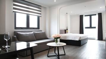 A modern hotel room with a sofa, a round table with a vase of flowers, a bed, and a large window.