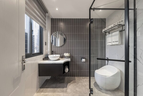 A modern bathroom with a large window, a round mirror, a white sink, and a glass-enclosed shower area.