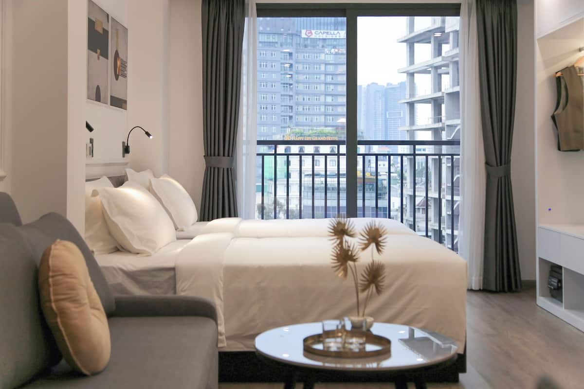 Une chambre d’hôtel moderne, équipée d’un lit, d’un canapé, d’une petite table avec un vase, et offrant une vue sur la ville grâce à de grandes fenêtres.
