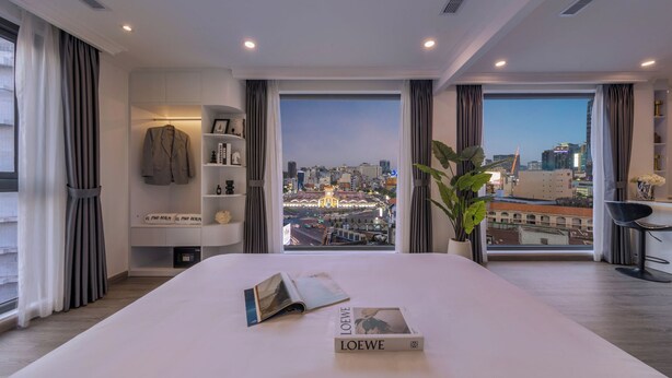A modern hotel room with a large bed, a city view, a bookshelf, and a desk with a chair. - CityHouse - El Pino Realm at Metro Ben Thanh Market (Ho Chi Minh City)