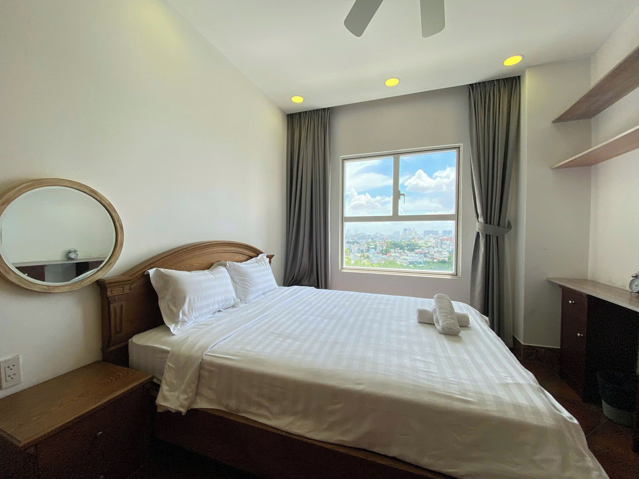 Habitación de hotel con una cama grande, un espejo redondo, una ventana con vistas a la ciudad y un escritorio con un reloj.