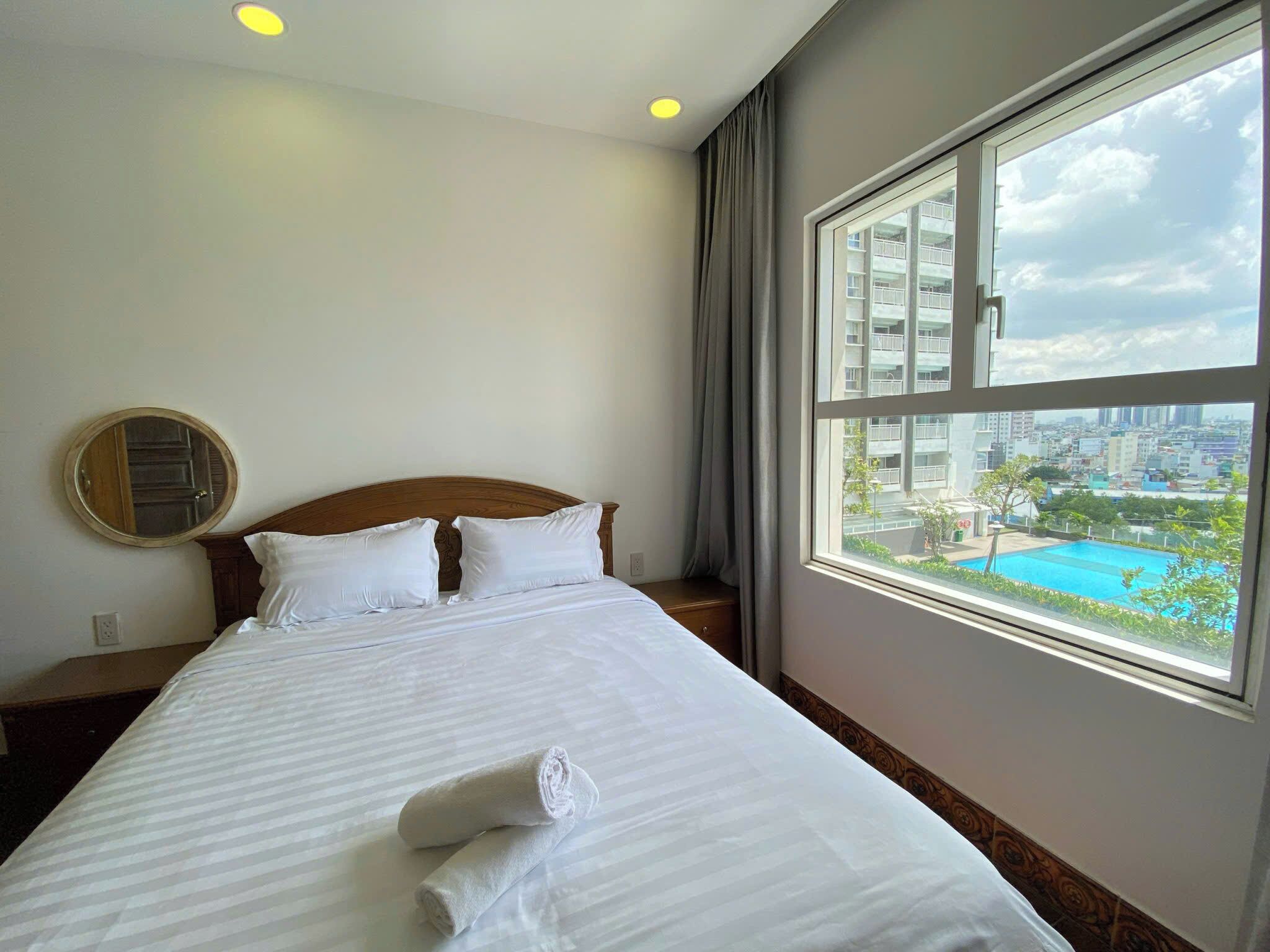 A hotel room with a large bed, a round mirror, a window with a city view, and a neatly made bed with towels.