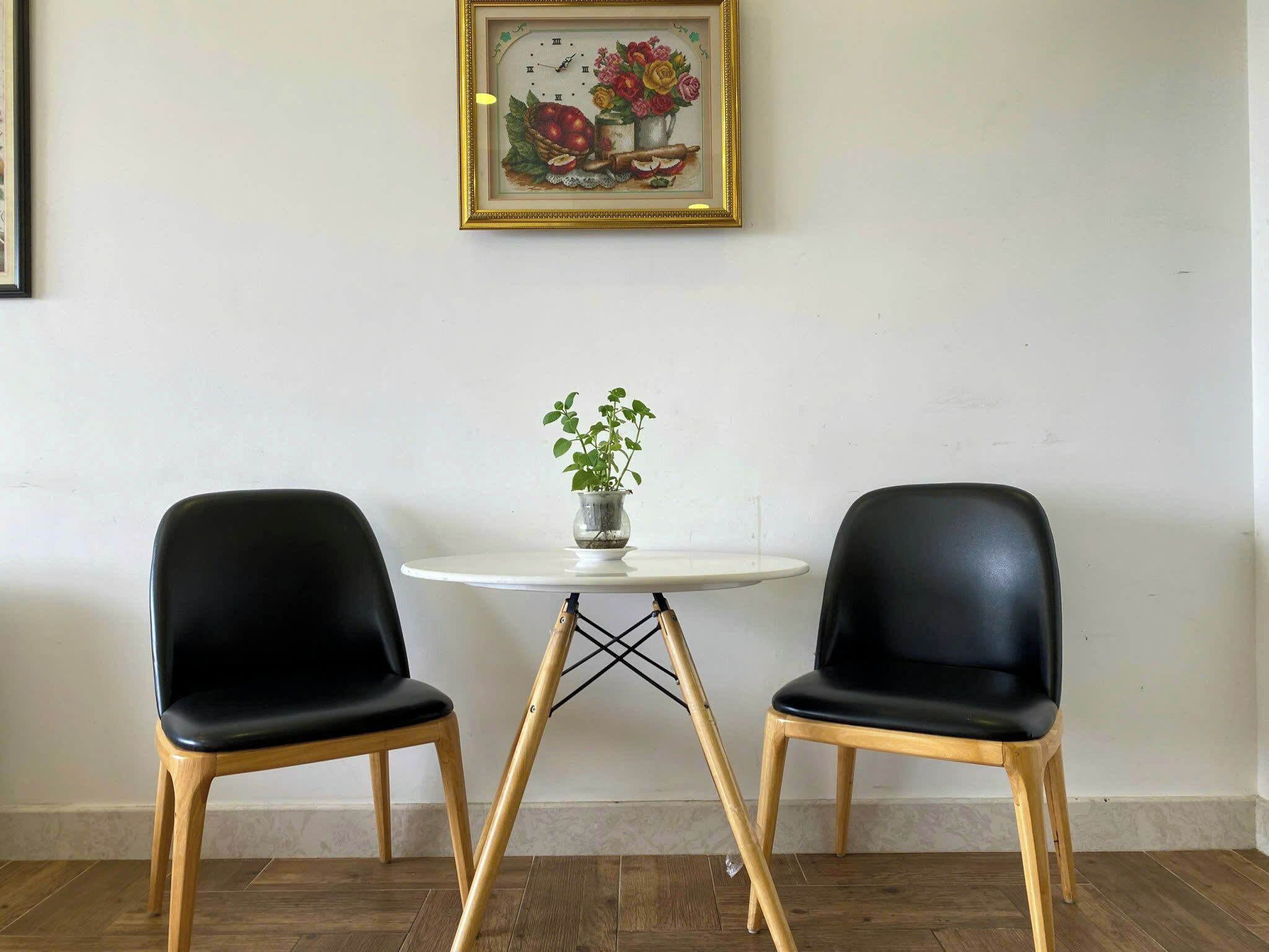 A room with two black chairs, a small round table with a potted plant, and a framed picture of flowers and apples on the wall.