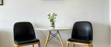 Een kamer met twee zwarte stoelen, een kleine ronde tafel met een plant in pot, en een lijst met bloemen en appels aan de muur.