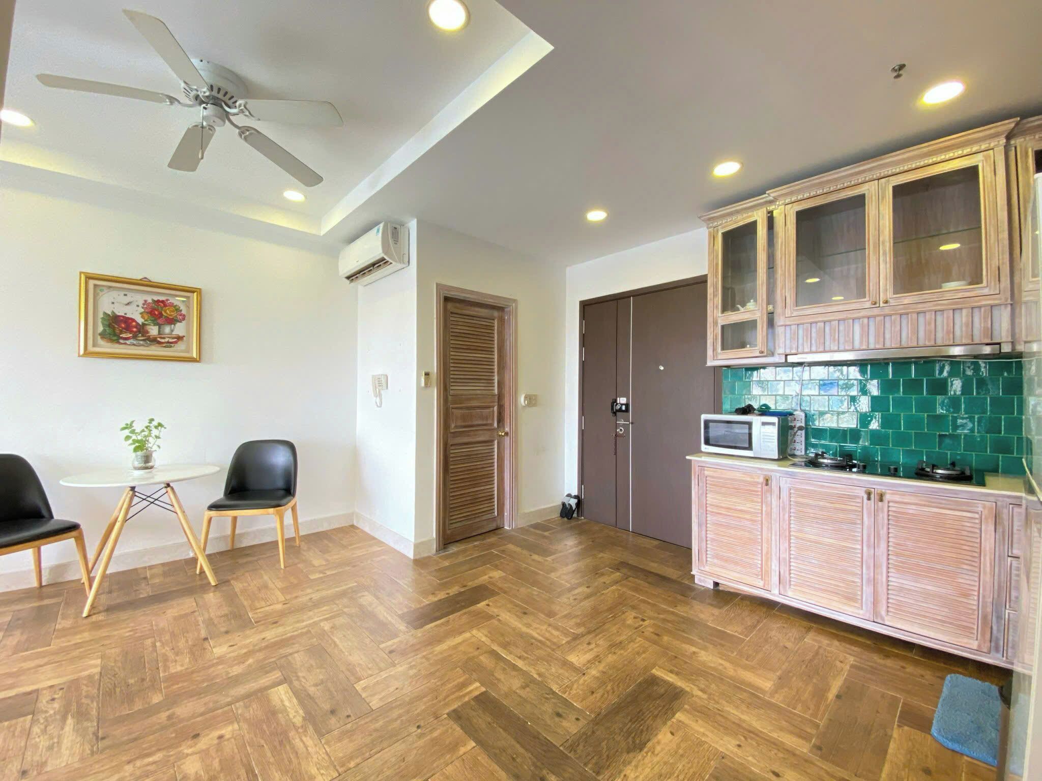 A room with a herringbone wooden floor, a ceiling fan, a dining area with two chairs and a small table, a kitchen with wooden cabinets and a microwave, and a door with a handle.