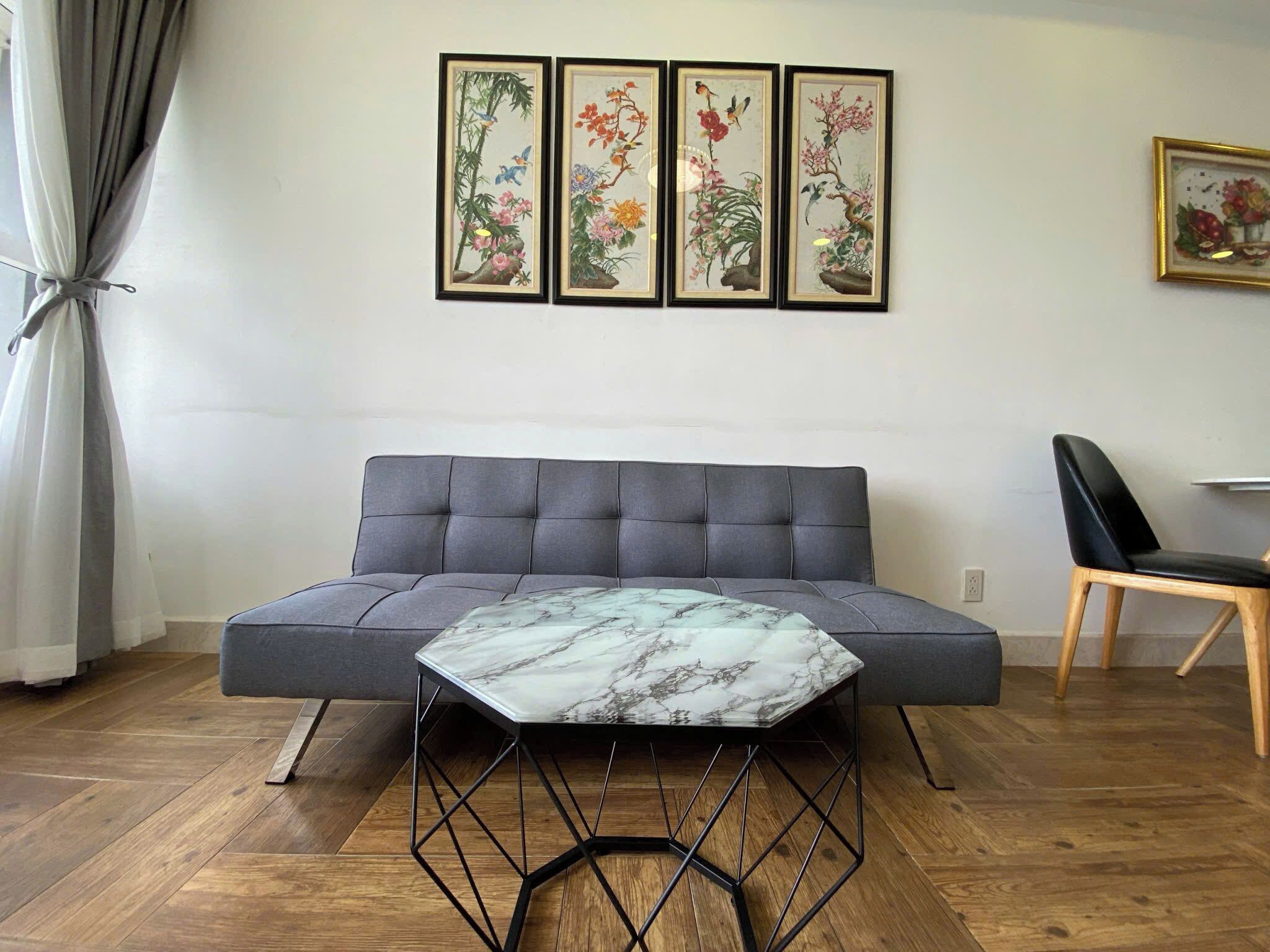 A living room with a grey sofa, a marble coffee table, and wooden flooring. There are three framed artworks on the wall and a chair to the right.